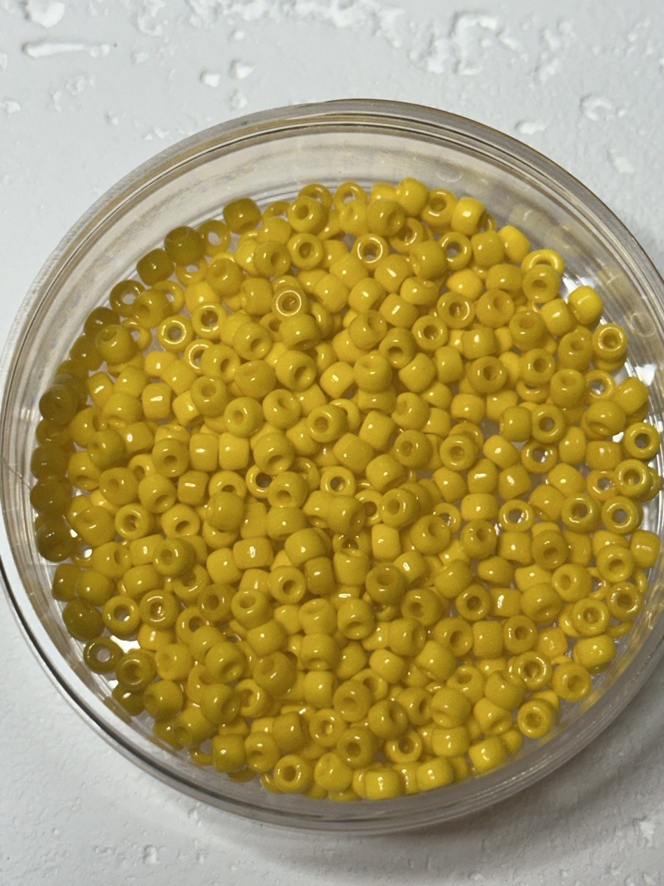 Yellow seed beads in a clear round container on a light textured background.