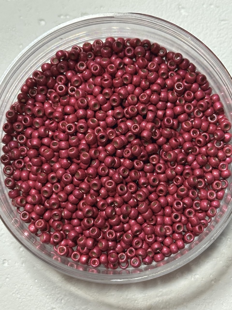 A top-down view of small, round, dark pinkish-red seed beads inside a clear plastic container, set against a light gray textured background.