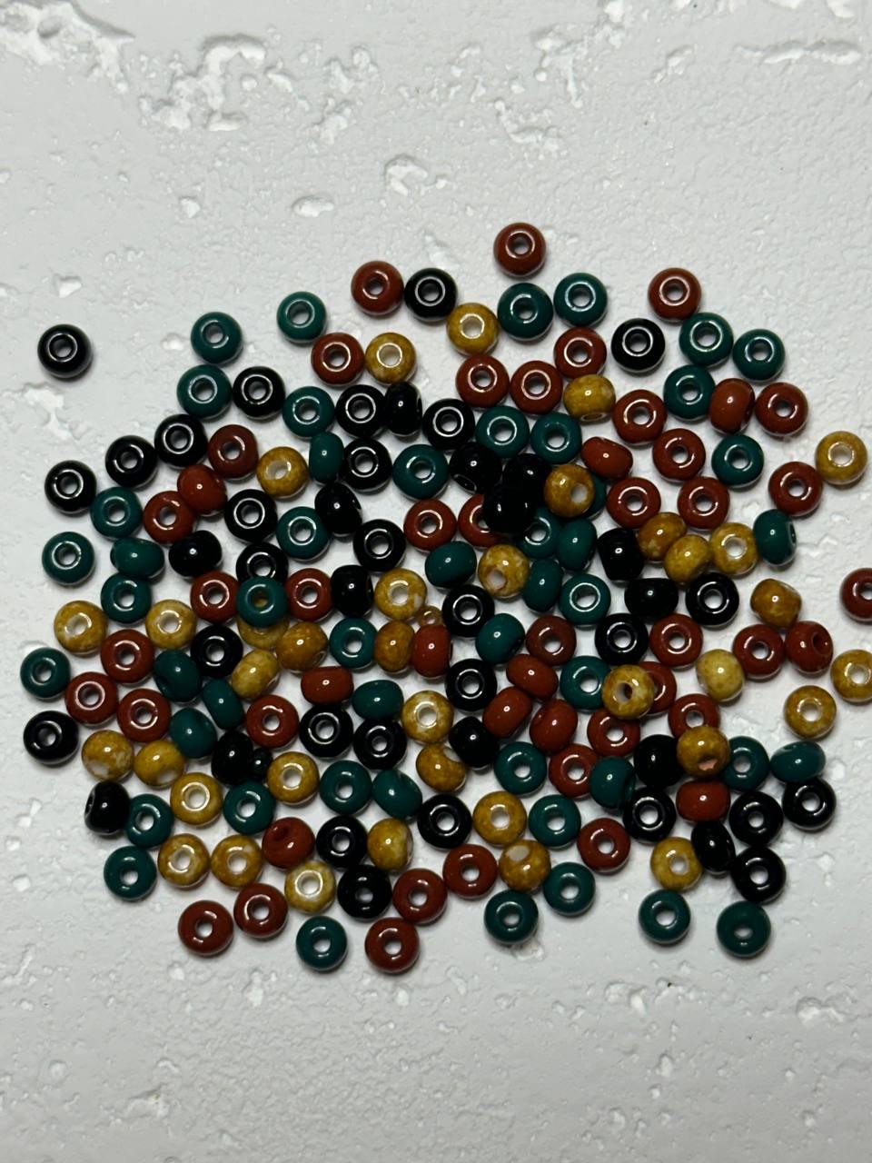 A pile of small round glass beads in multiple colors, including dark green, black, rust red, and mustard yellow, scattered on a textured white surface.