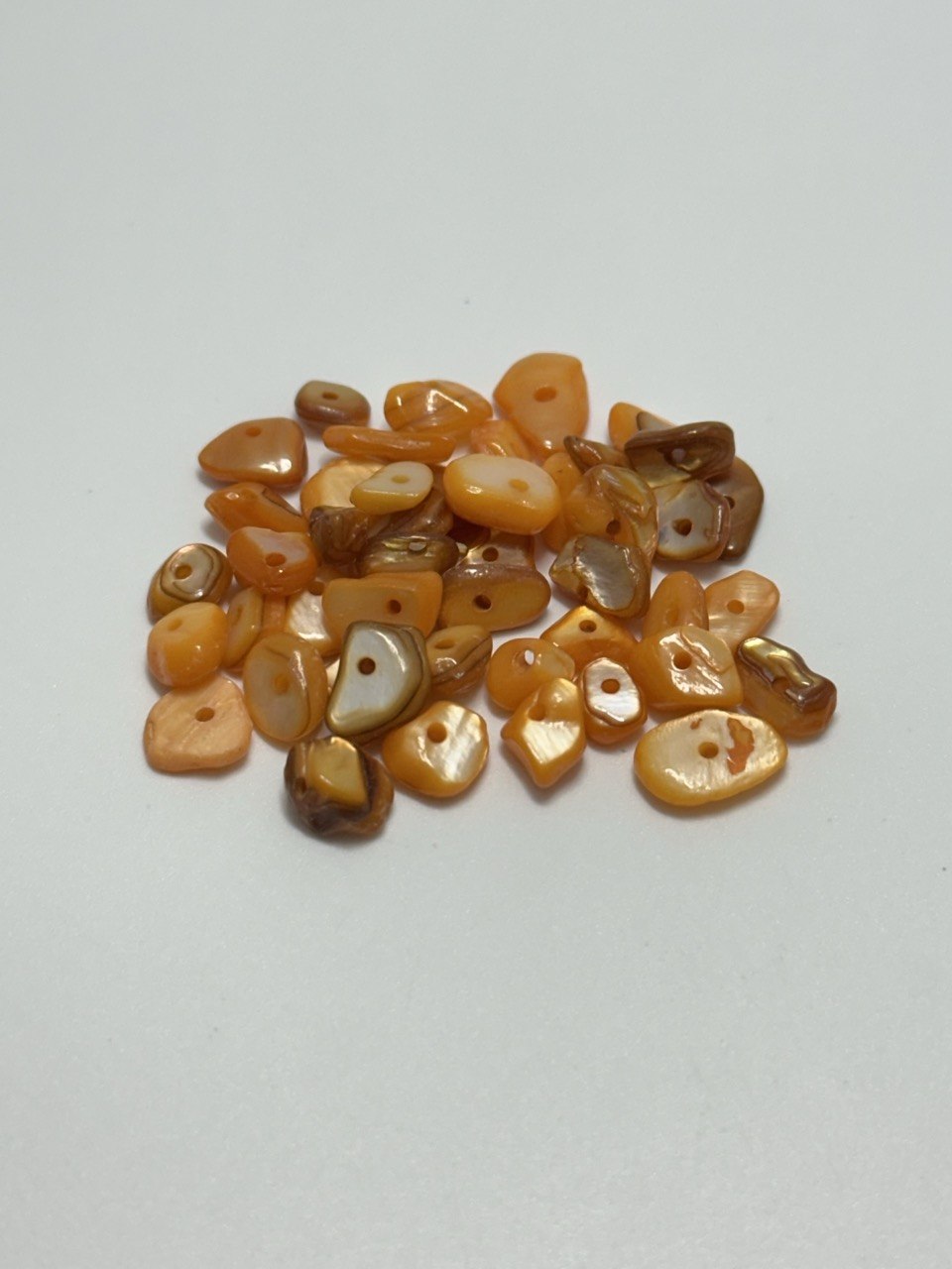 A small pile of irregular, tumbled dyed orange mother of pearl beads with pre-drilled holes, displayed on a plain light gray background.