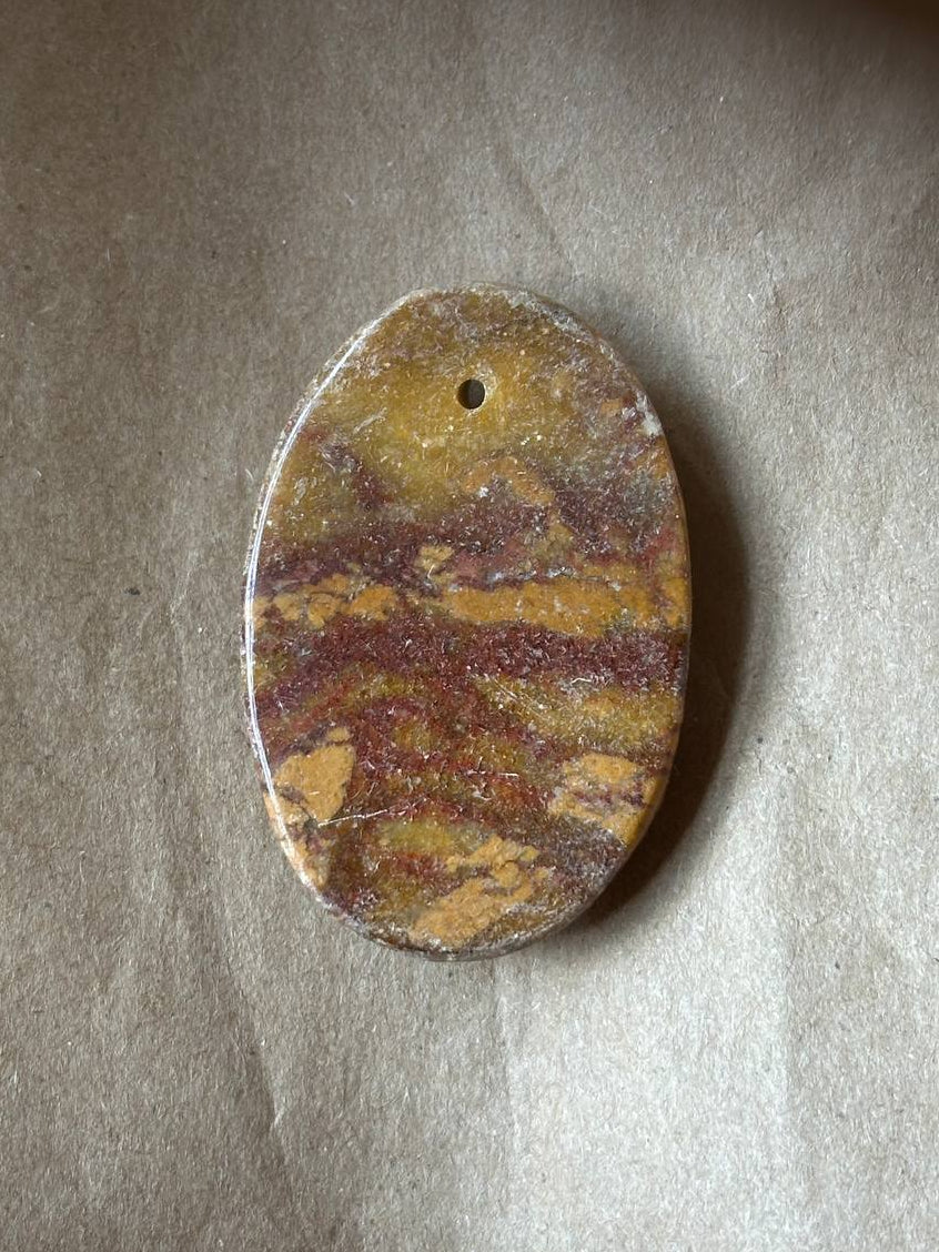 A polished oval stone pendant with a natural marbled pattern of yellow, brown, and orange, featuring a small hole at the top for hanging, displayed on a textured beige surface.