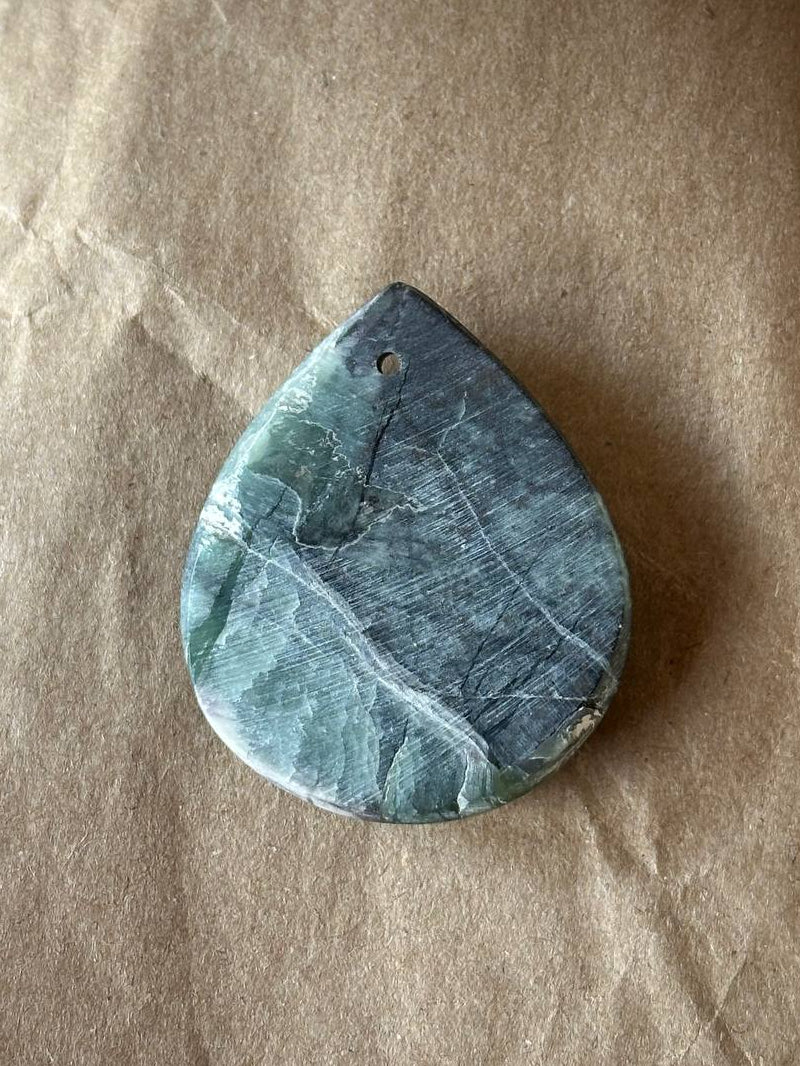 A polished teardrop-shaped gemstone pendant with a dark teal and gray marbled pattern, featuring a small pre-drilled hole at the top, displayed on a textured brown paper background.