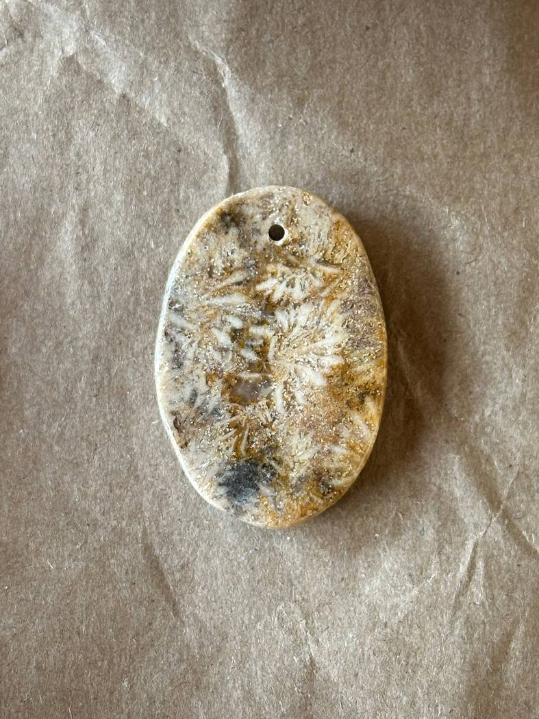 An oval polished stone pendant with a natural marbled pattern in beige, brown, and gray, featuring a small hole at the top, displayed on a textured brown paper background.