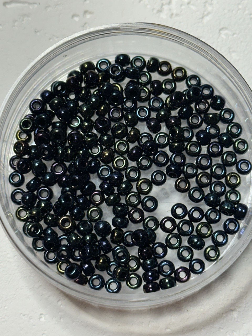 A top-down view of small iridescent dark beads inside a clear round plastic container, set on a light textured surface.