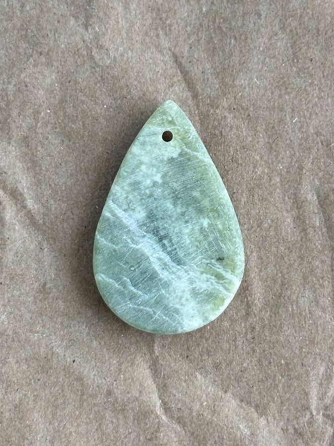 A teardrop-shaped light green stone pendant with natural white veining and a small hole at the top, placed on a textured brown paper background.