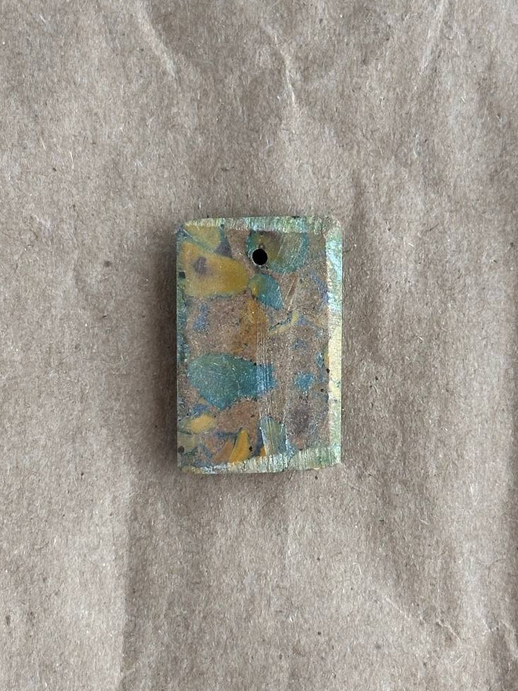 A rectangular stone pendant with a hole at the top, featuring a marbled pattern of blue, yellow, and gray, resting on a textured gray surface.