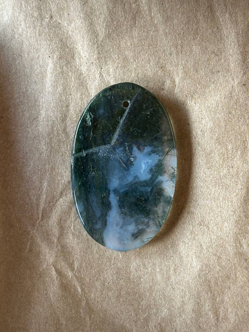 An oval polished stone pendant with a dark green and blue marbled pattern and white cloudy areas, resting on a textured beige paper background.