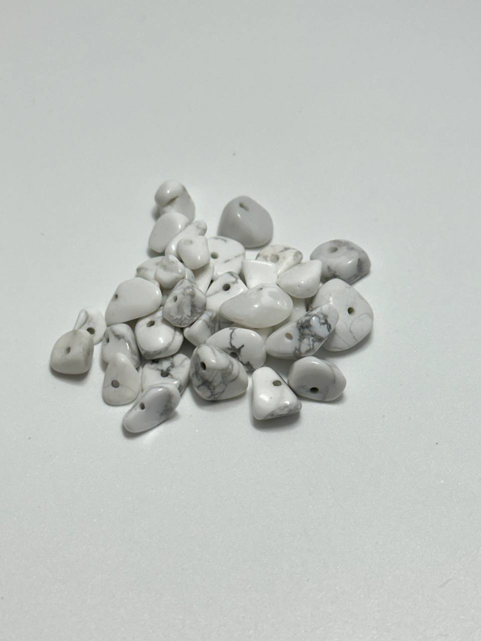 A pile of small, polished white stone chips with gray veining and pre-drilled holes, displayed on a plain light gray background.