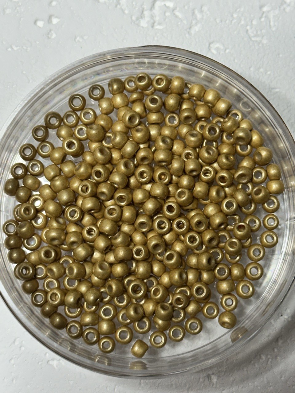 Metallic gold seed beads with a polished finish, piled inside a clear plastic round container on a textured light gray background.