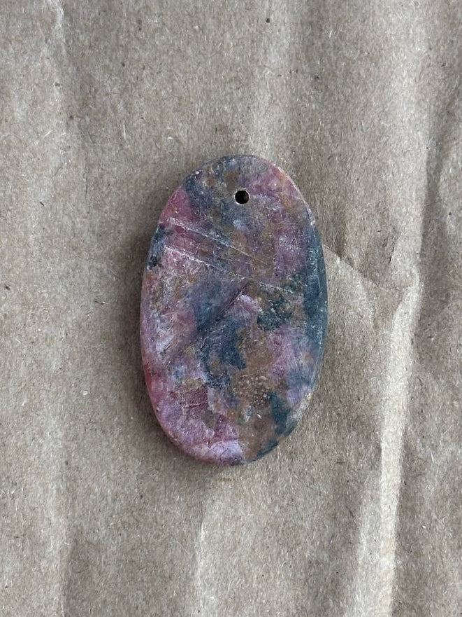 An oval-shaped cabochon gemstone pendant with a marbled pattern of pink, purple, and blue, featuring a small hole at the top for hanging, placed on a textured brown paper background.