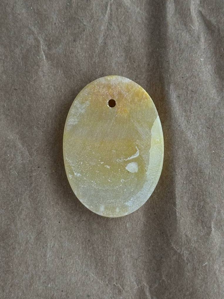 An oval-shaped pale yellow and white marbled stone pendant with a small hole at the top, displayed on a textured gray background.