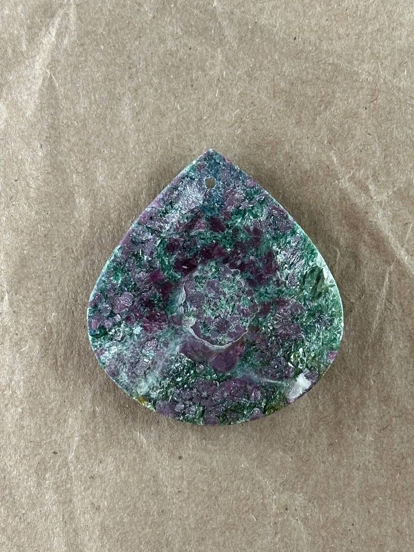 A teardrop-shaped gemstone cabochon with a marbled pattern of green, purple, and white, displayed on a textured brown paper background.