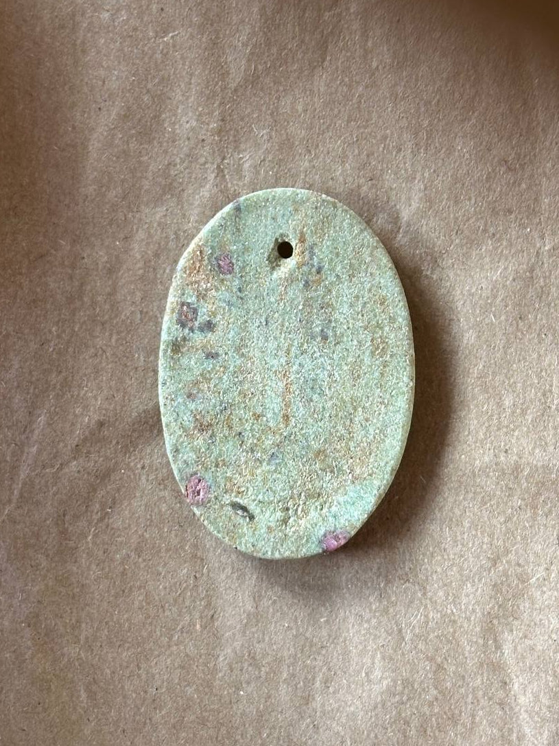 An oval-shaped pendant with a weathered, patinated surface showing light green and beige tones, featuring a small hole at the top for hanging, placed on a textured brown paper background.