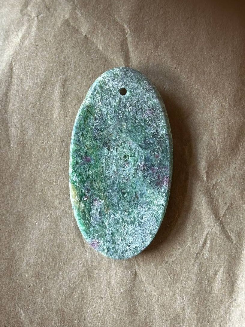 An oval-shaped natural stone pendant with a textured, speckled pattern of green, blue, and purple, featuring a small hole at the top for hanging, displayed on a brown kraft paper background.