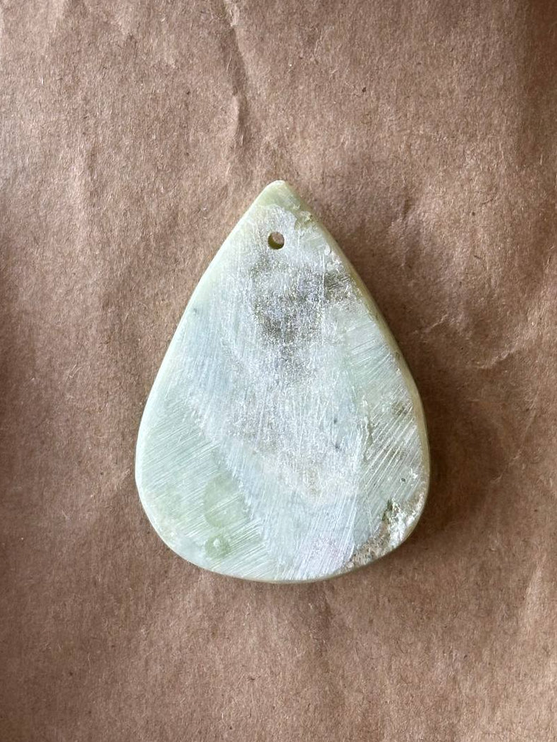 A teardrop-shaped light green stone pendant with a small hole at the top, resting on a brown textured paper background.