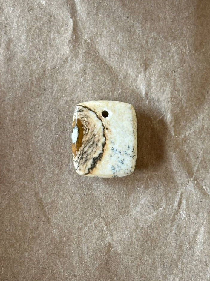 A square natural stone bead with a hole on one side, featuring a textured pattern of cream, brown, and light blue, displayed on a sheet of brown kraft paper.