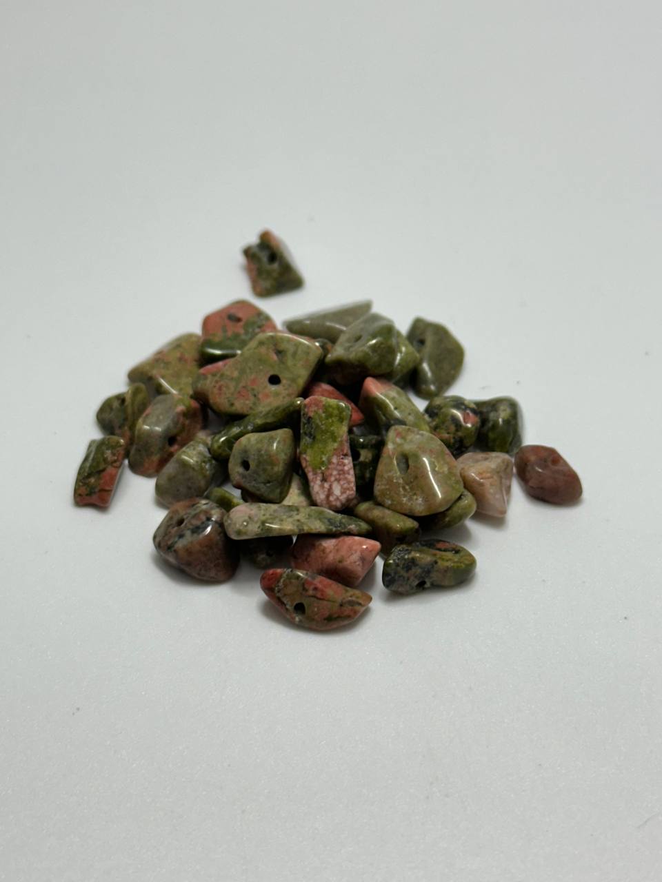 A small pile of polished tumbled stone beads or pendants in mixed earthy colors, including olive green, reddish pink, brown, and gray, arranged on a plain light background.