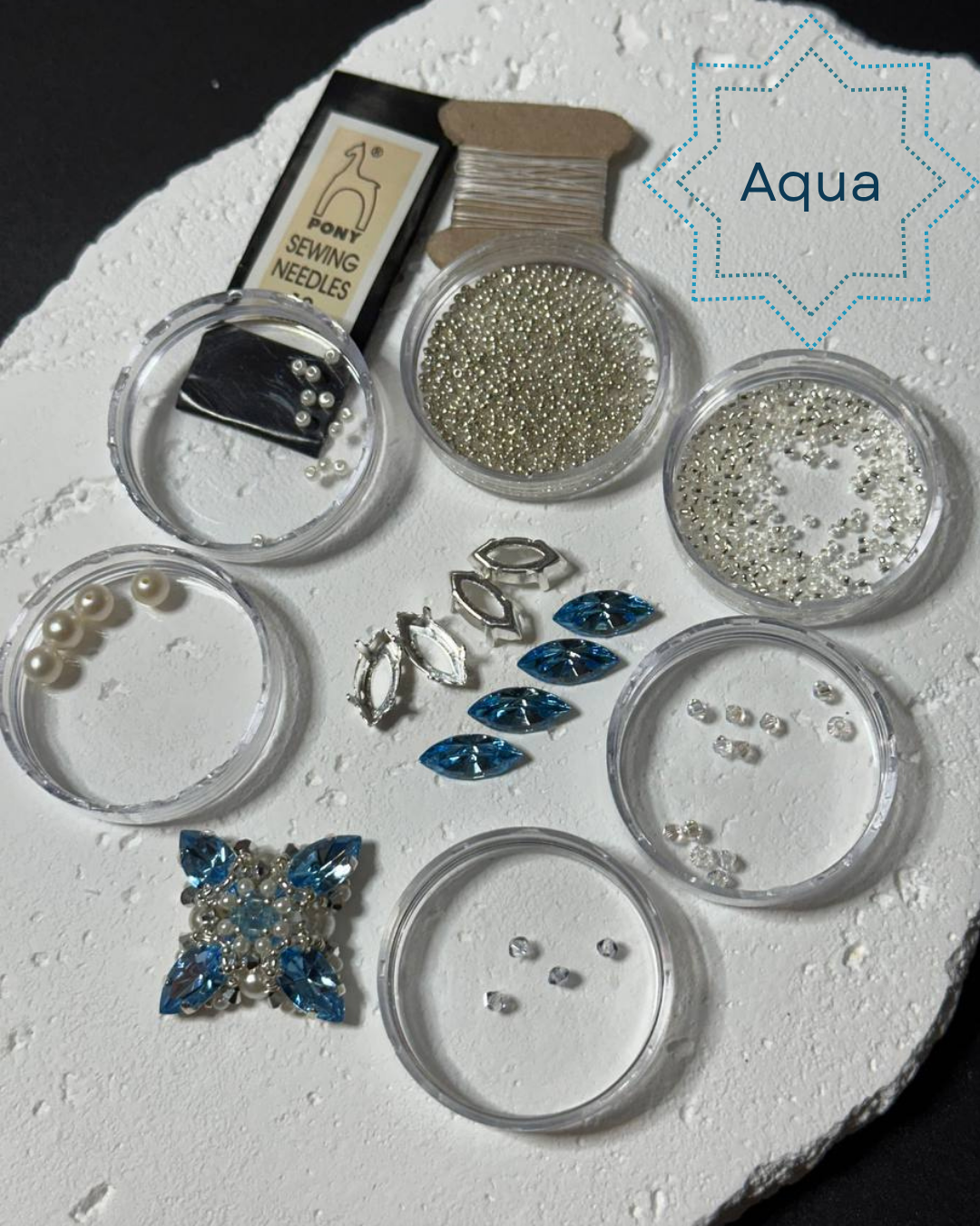 A jewelry embellishment kit labeled 'Aqua' containing various small beads, rhinestones, pearls, sewing needles, thread, and a blue floral applique on a white textured background.