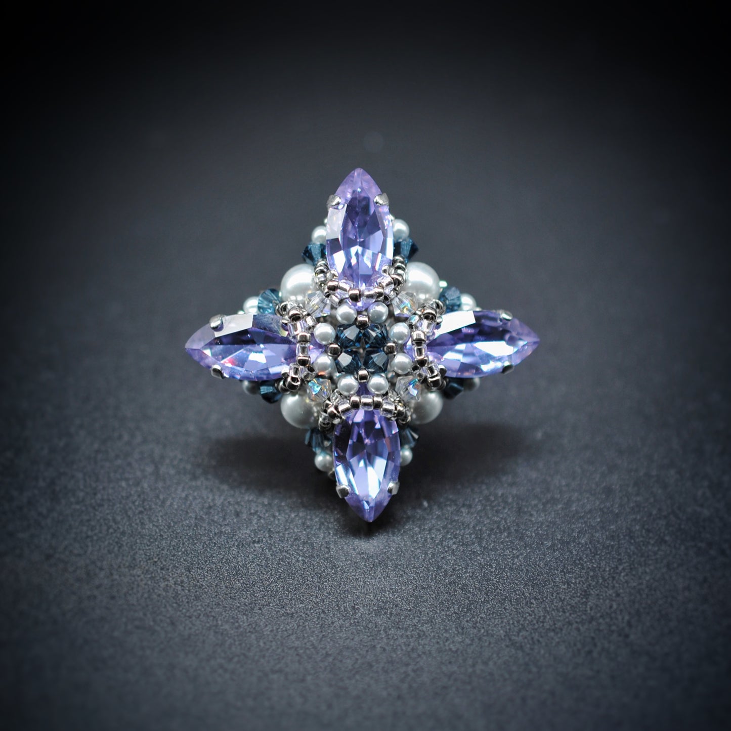 A decorative four-pointed crystal brooch or pin featuring light purple marquise-cut crystals, small white pearls, and blue faceted accents, photographed on a dark gray background.
