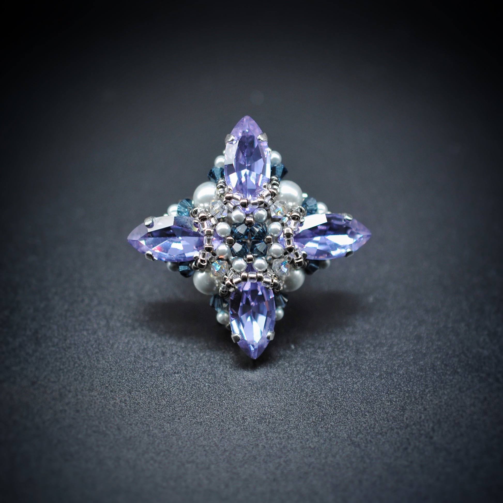 A decorative four-pointed crystal brooch or pin featuring light purple marquise-cut crystals, small white pearls, and blue faceted accents, photographed on a dark gray background.