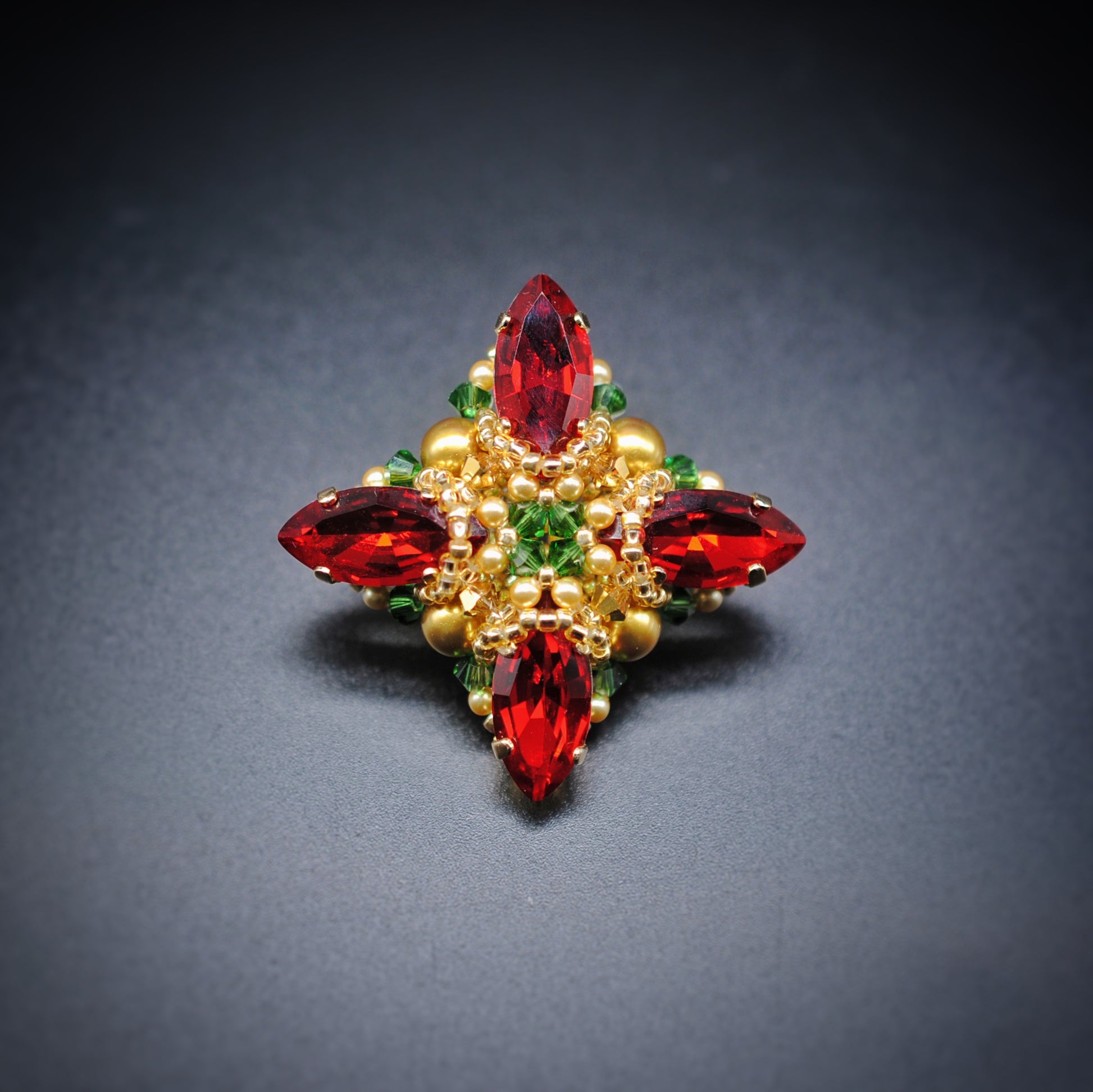 A vintage-style floral brooch featuring four marquise-cut red crystals, accented with gold beads and small green crystals, set against a dark gray background.