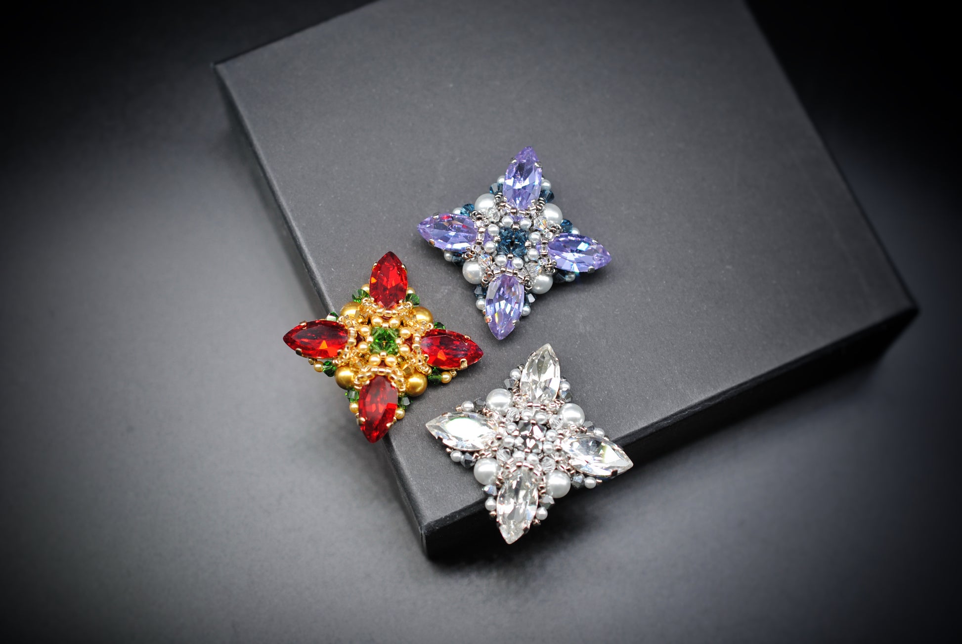 Three star-shaped rhinestone brooches in red, blue, and silver, displayed on a black rectangular box against a dark gray background.