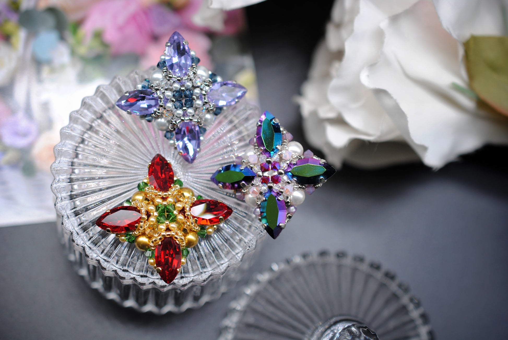 Three decorative crystal brooches displayed on a clear glass dish, with a blurred floral background.