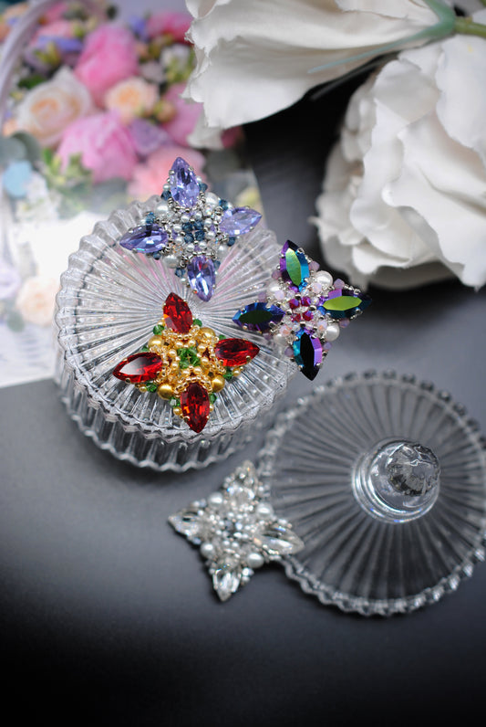 A set of clear glass keepsake boxes with matching lids, decorated with colorful crystal star-shaped brooches, displayed on a dark surface with white and pink flowers in the background.