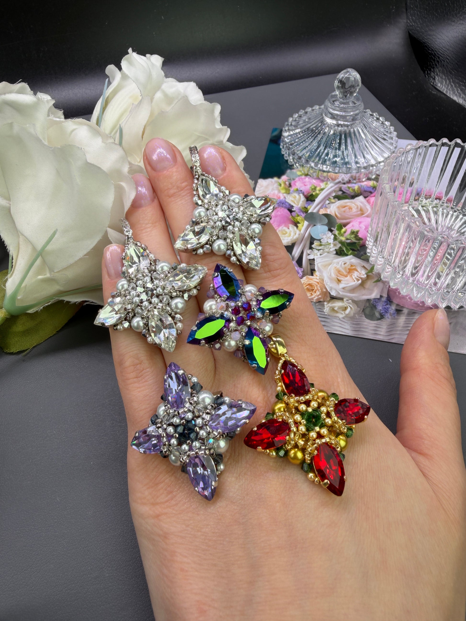 A hand holds four colorful star-shaped rhinestone brooches or pendants with silver, purple, green, and red stones, set against a styled background with white roses and glassware.