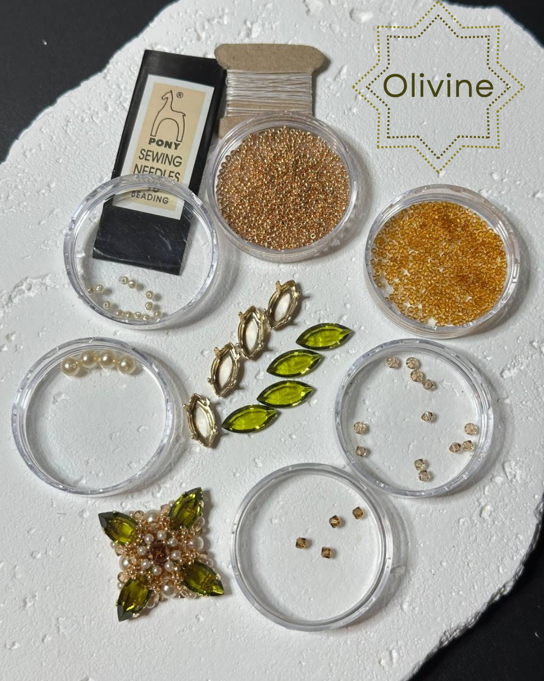 Olive green marquise-cut seed beads displayed with other sewing and embellishment supplies on a white textured surface. The image includes a package of Pony Sewing Needles, gold and bronze seed beads, pearl beads, a star-shaped embellishment, and a brand label reading 'Olivine'.