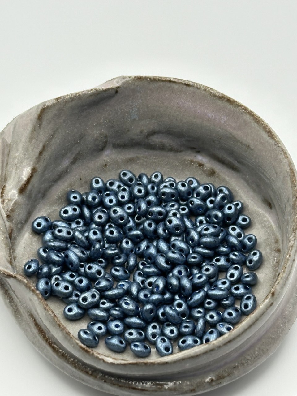 Ceramic bowl filled with blue superduo beads on a white background