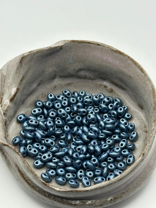 Ceramic bowl filled with blue superduo beads on a white background