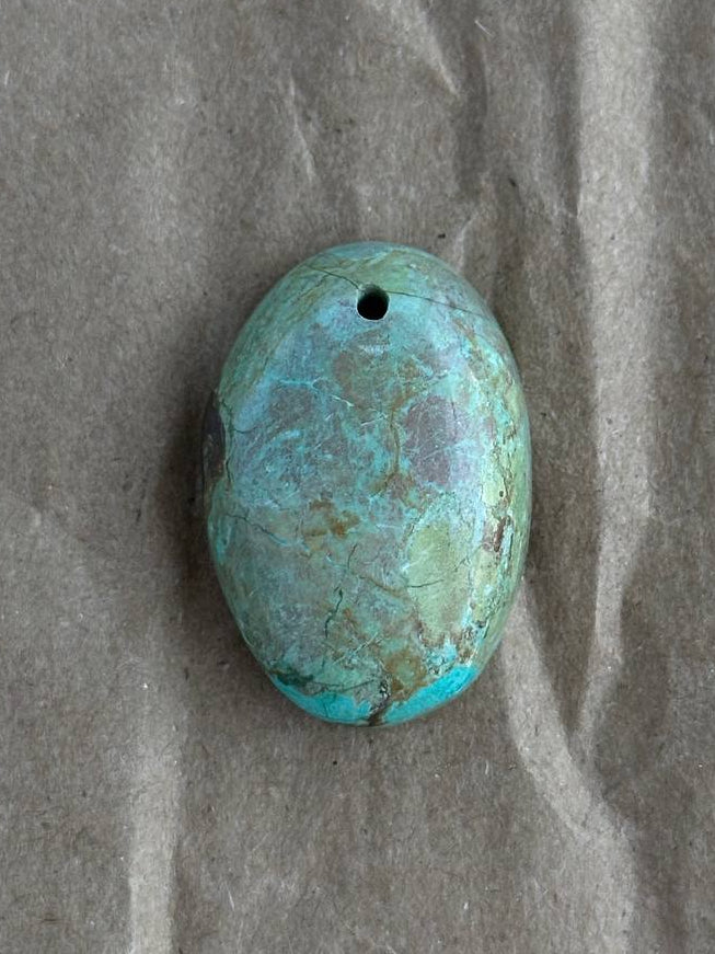 An oval chrysocolla stone bead with a hole at the top, resting on a textured brown paper background.