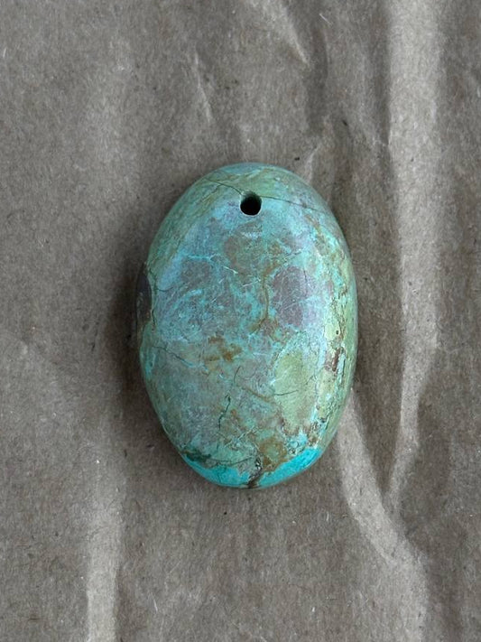 An oval chrysocolla stone bead with a hole at the top, resting on a textured brown paper background.