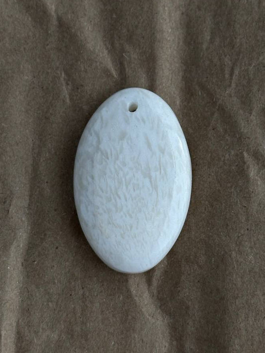 An oval white stone pendant with a small hole at the top, resting on a brown textured paper background.