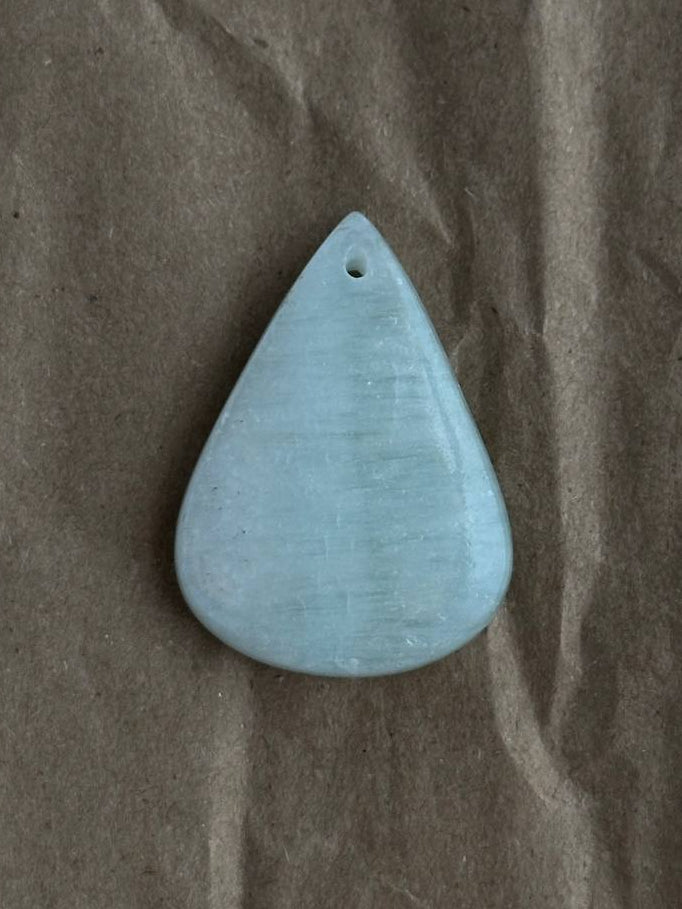 A smooth, teardrop-shaped light blue stone pendant with a small hole at the top, resting on a textured brown paper background.