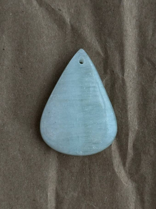 A smooth, teardrop-shaped light blue stone pendant with a small hole at the top, resting on a textured brown paper background.