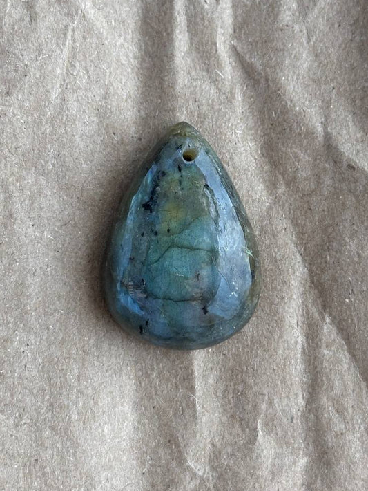 A teardrop-shaped labradorite stone pendant with a small hole at the top, resting on a textured beige paper background.