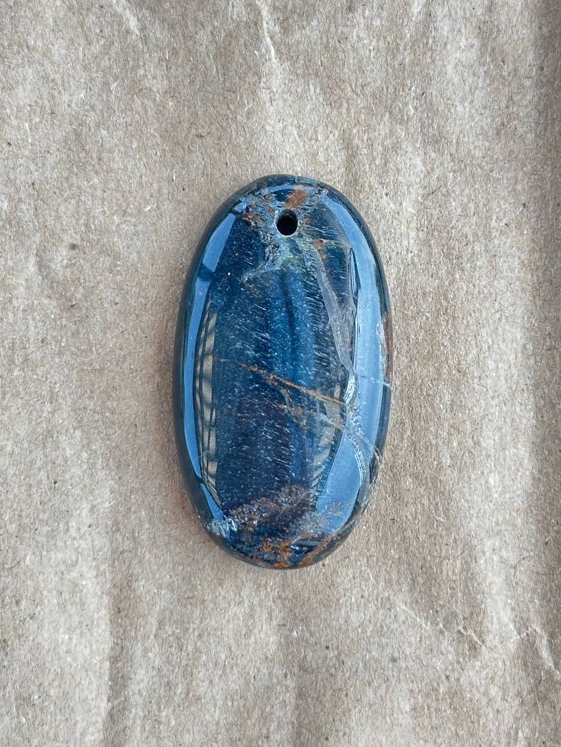 A polished blue pietersite pendant with natural brown and white inclusions, displayed on a textured beige fabric background.