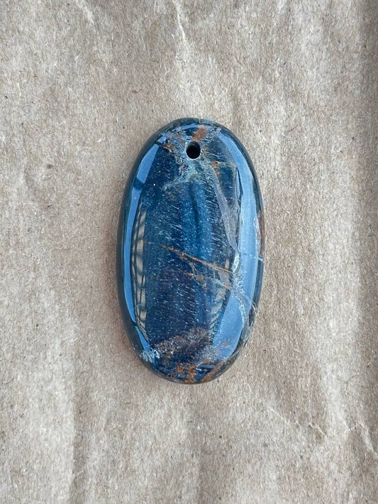 A polished blue pietersite pendant with natural brown and white inclusions, displayed on a textured beige fabric background.