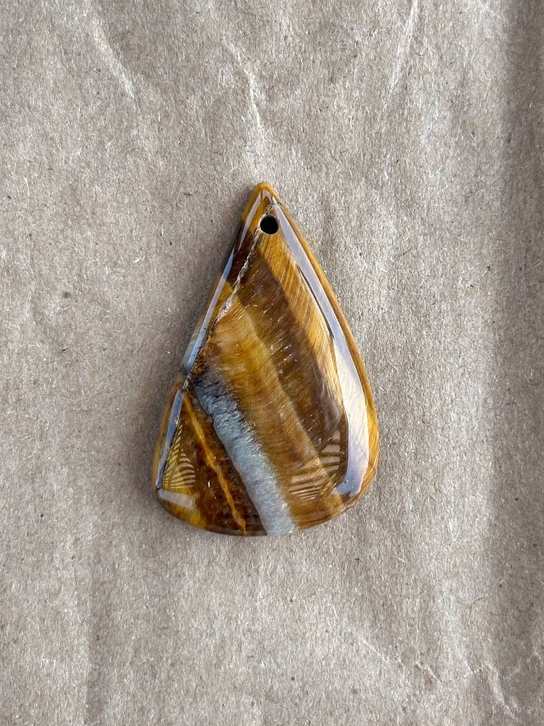 A teardrop-shaped pendant with a natural tiger's eye pattern, featuring brown, gold, and gray banded bands, resting on a textured beige surface.