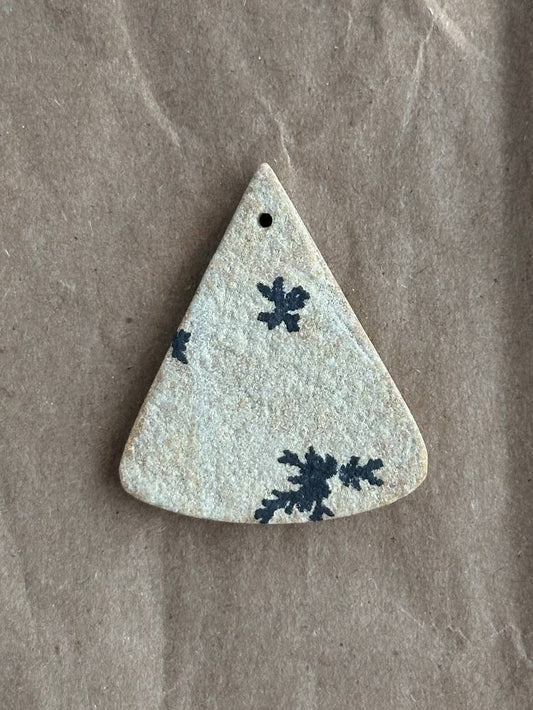 A triangular druzy pendant with a small hole at the top, featuring a dark blue snowflake pattern on a textured off-white glaze, displayed on a brown kraft paper background.