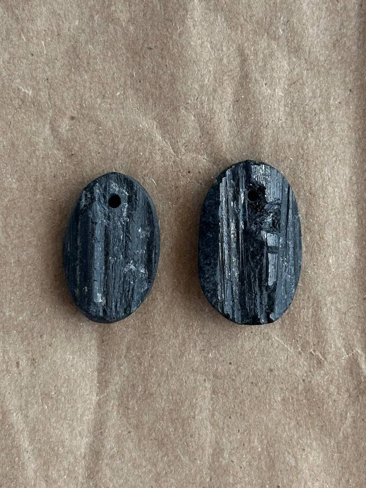 Two small oval dark gray stone beads with a woodgrain texture and a hole at the top, displayed on a brown kraft paper background.