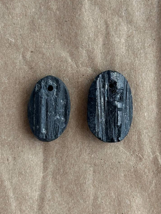 Two small oval dark gray stone beads with a woodgrain texture and a hole at the top, displayed on a brown kraft paper background.