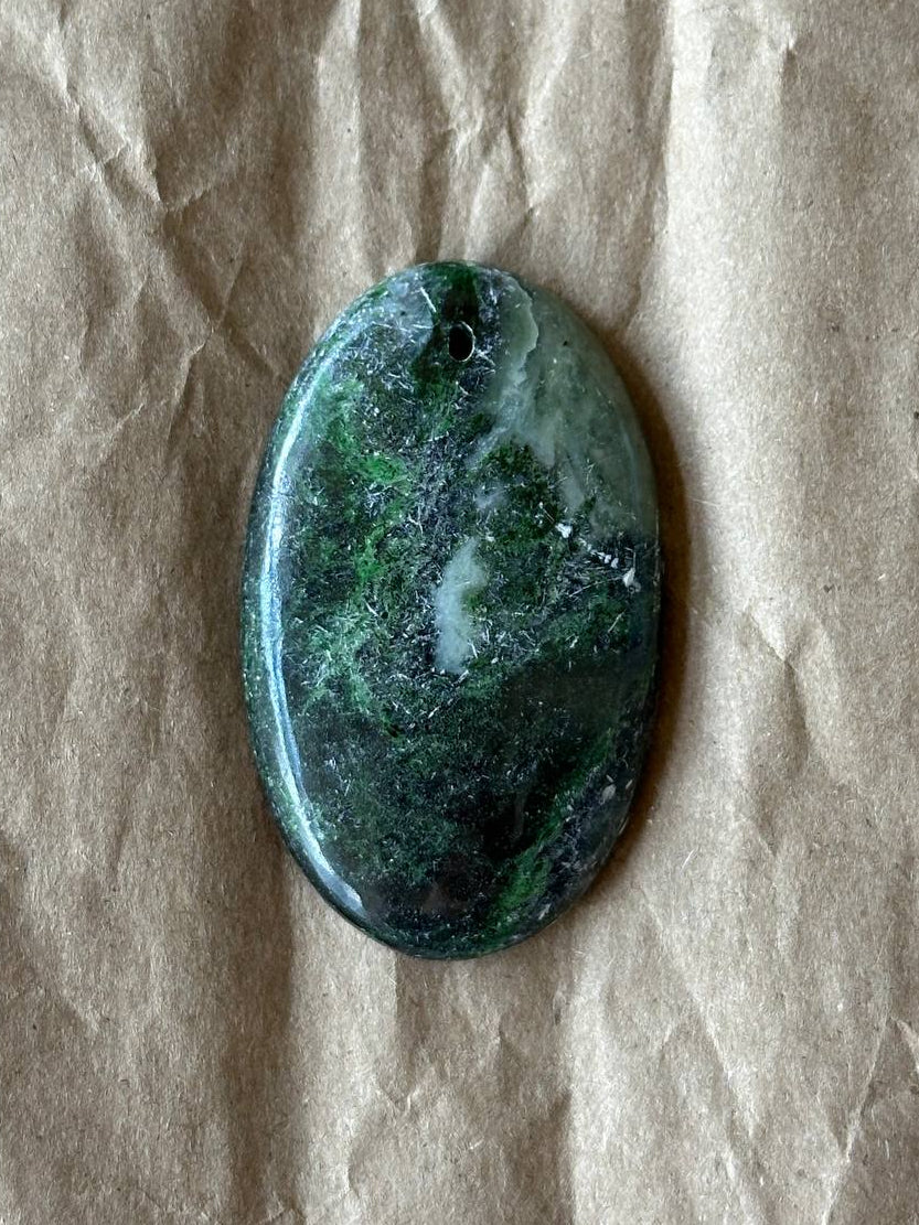 An oval green stone pendant with natural marbled patterns, featuring a small hole at the top for hanging, displayed on a textured beige paper background.
