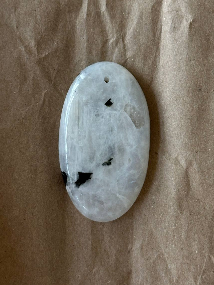 An oval-shaped white stone pendant with natural dark inclusions and a small hole at the top, resting on a textured brown paper background.