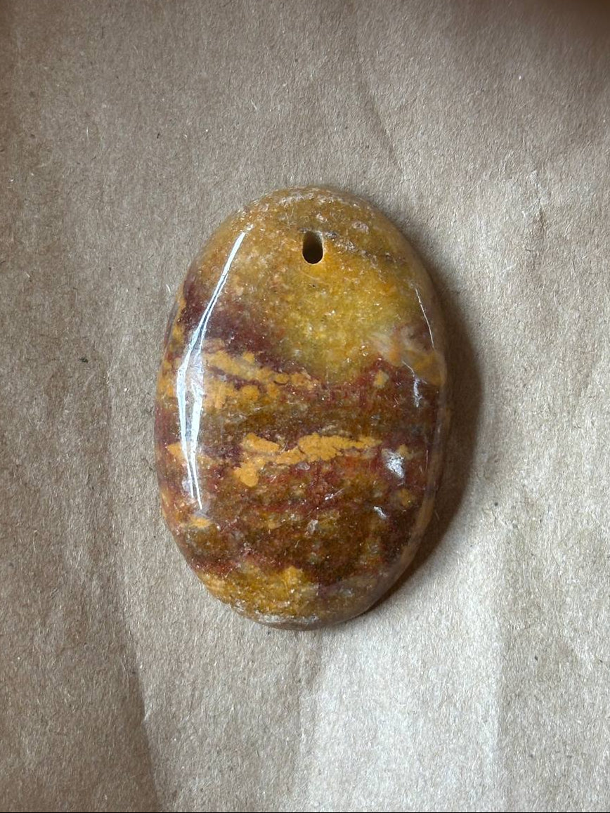 A polished oval stone pendant with a natural marbled pattern of yellow, brown, and orange, featuring a small hole at the top for hanging, displayed on a textured beige surface.