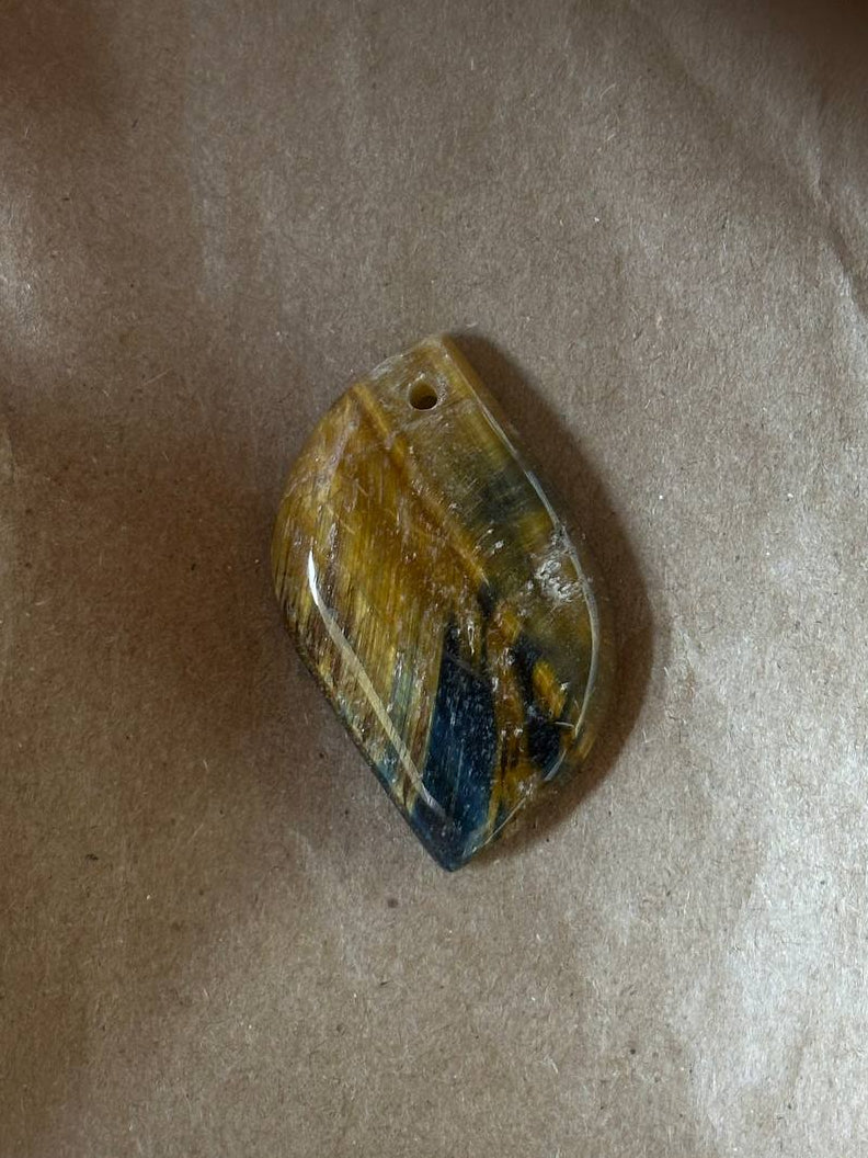 A small polished stone pendant with a hole at the top, showing natural marbled patterns of yellow, brown, and dark blue, resting on a sheet of brown paper.