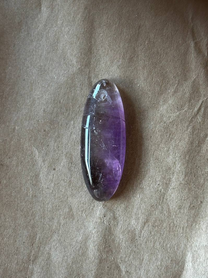 A polished oval amethyst gemstone with a gradient of purple and gray tones, resting on a textured beige paper background.