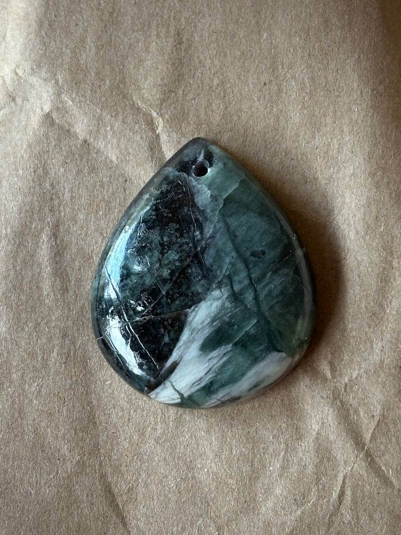 A polished teardrop-shaped gemstone pendant with a dark green, black, and white marbled pattern, resting on a textured brown paper background.